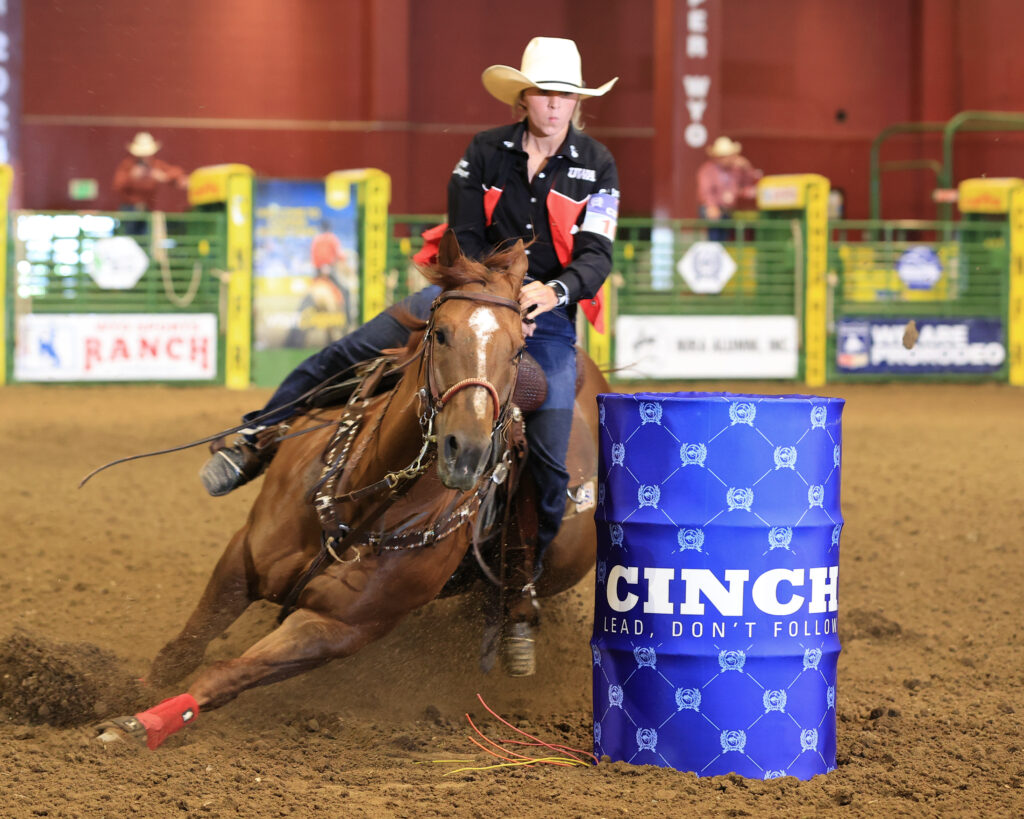 Barrel racer in a white cowboy hat rounds a blue CINCH barrel as her sorrel horse digs low into the dirt