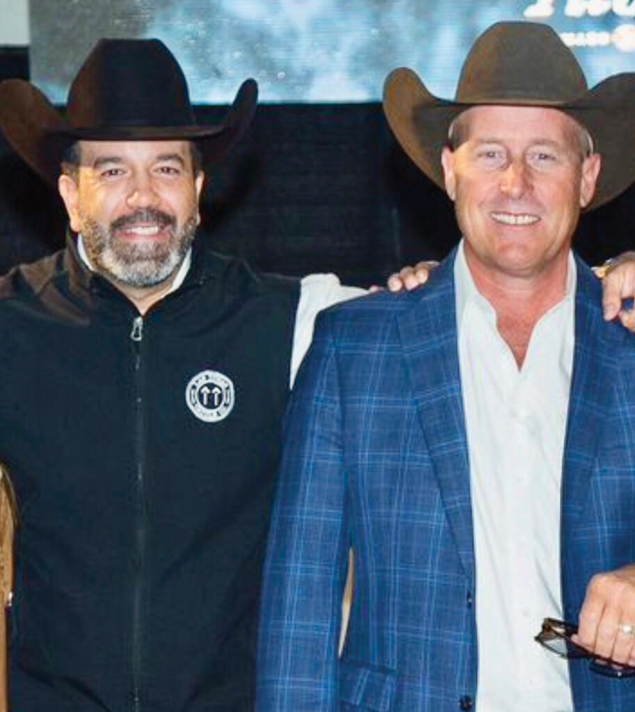 Two men in cowboy hats pose together at what appears to be a rodeo event, with one wearing a black jacket and the other in a blue plaid blazer