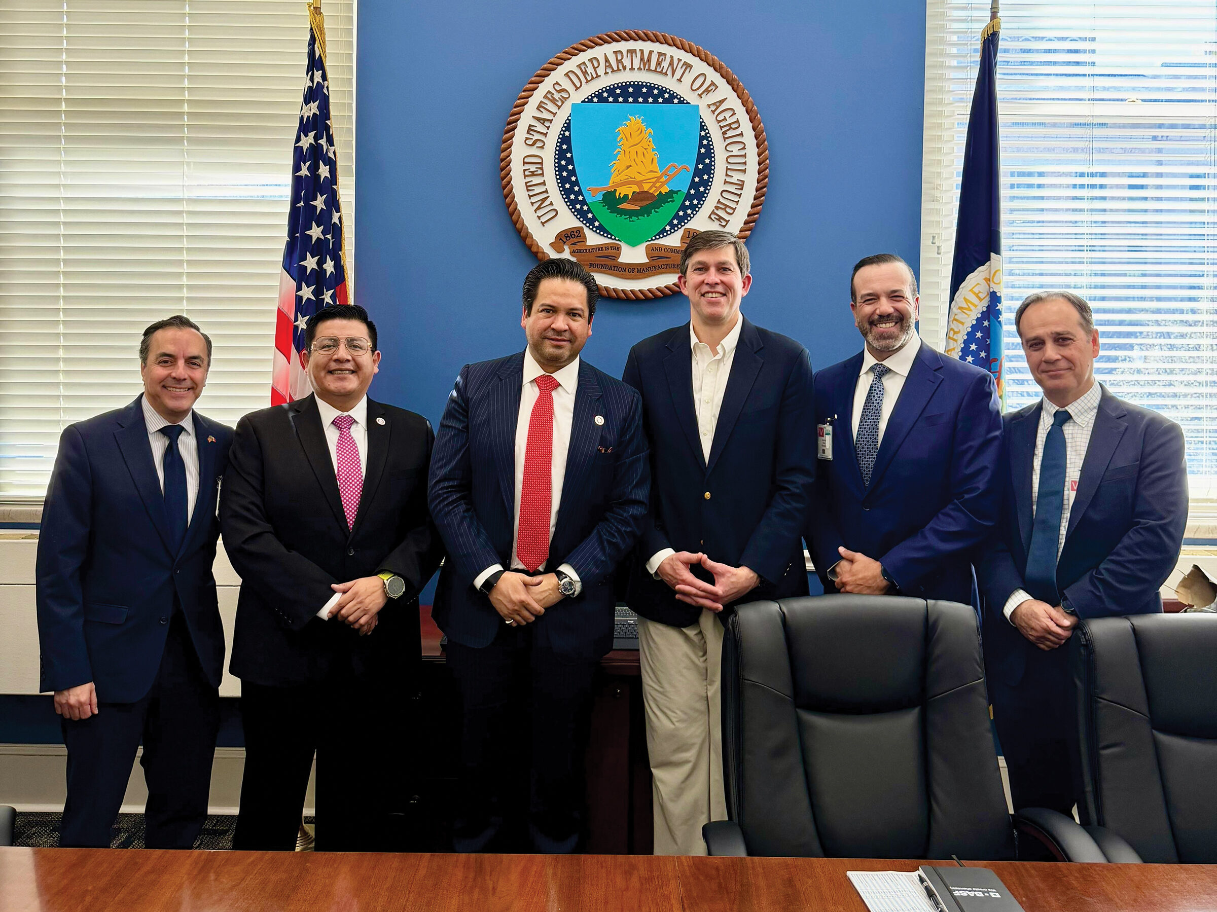 Six men in business attire stand together in front of the USDA Department of Agriculture seal in an official office setting