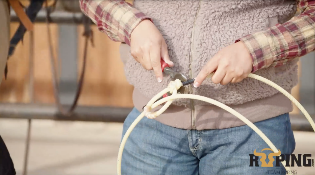 Close-up of a roper in a plaid shirt and fleece vest using red-handled pliers to work on the burner of a team roping rope