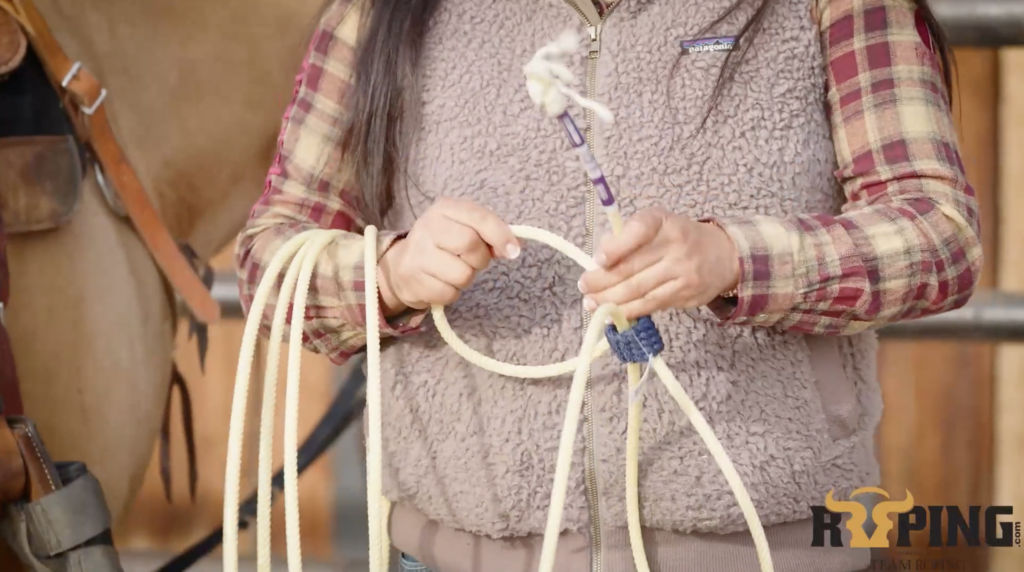 Woman in a plaid shirt and fleece vest holds a coiled team roping rope, demonstrating how to build a loop beside a horse