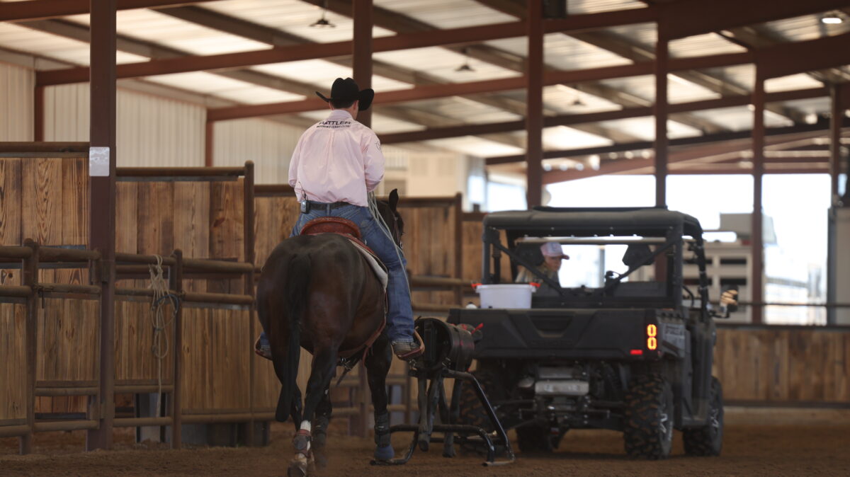 Cowboy in a black hat and pink shirt rides a dark horse toward a roping dummy and UTV inside a covered arena