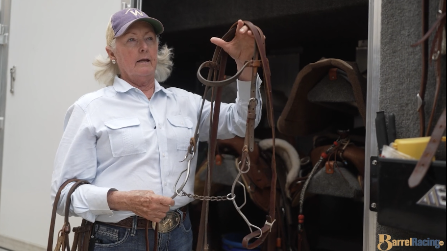 Woman in a ball cap and light button-up shirt holds up a bridle with a shanked bit inside a horse trailer tack room