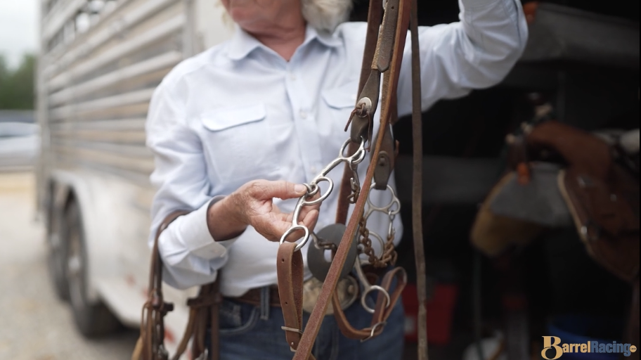 Man in a light blue shirt holds up a horse bit and headstall while examining the bridle beside a stock trailer