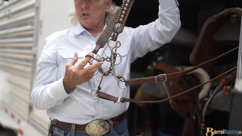 Woman in a light blue western shirt and trophy buckle holds up a leather headstall with a shanked curb bit and chain, explaining its parts beside a horse trailer