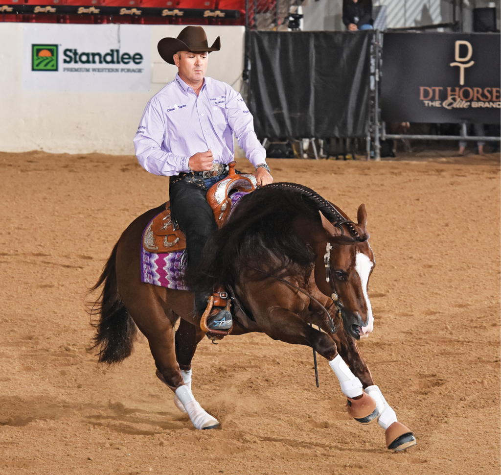 Reiner in a lavender shirt and black cowboy hat guides his bay horse through a sliding maneuver in an arena