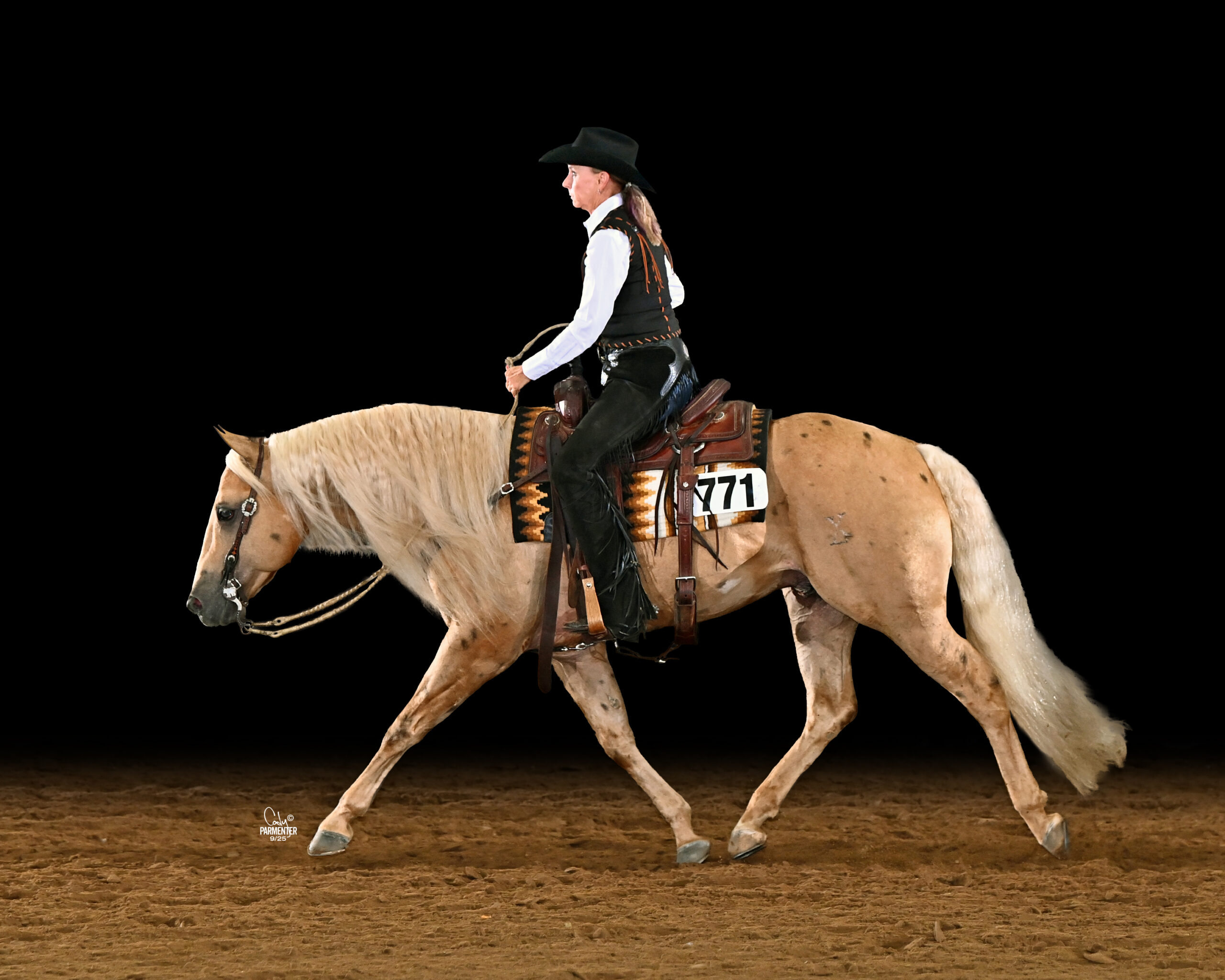 Western pleasure rider in black hat, vest and chaps jogs a palomino horse wearing number 771 across an arena
