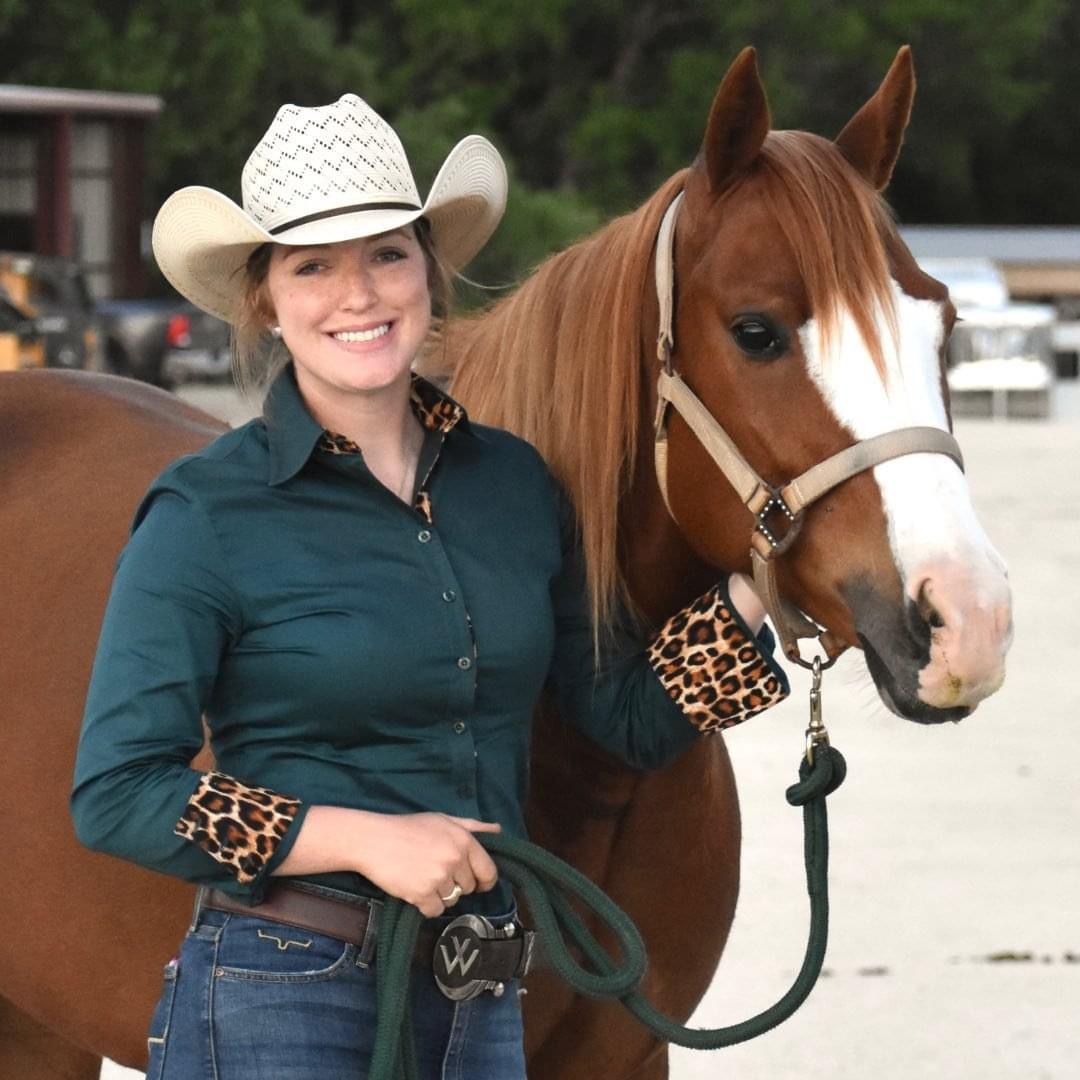 Woman in western attire with white cowboy hat posing with brown and white horse at rodeo arena