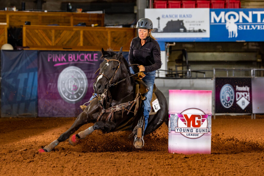 Barrel racer in a helmet rounds a Young Guns League barrel on a dark horse, dirt flying in an indoor arena