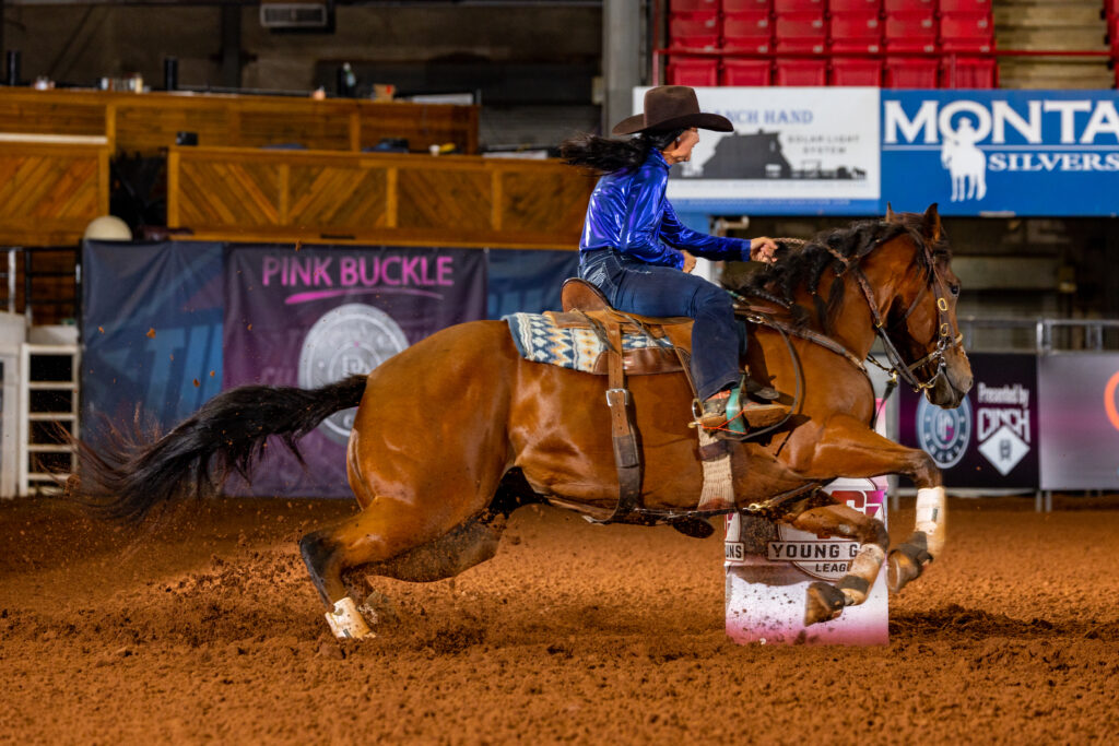 Barrel racer in a blue shirt and black cowboy hat turns a bay horse tight around a barrel, kicking up arena dirt