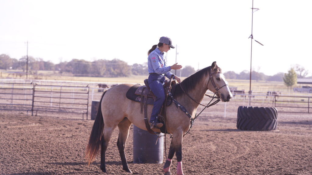 Rider in a ball cap sits on a buckskin horse beside a barrel in a dirt arena, gesturing with one hand