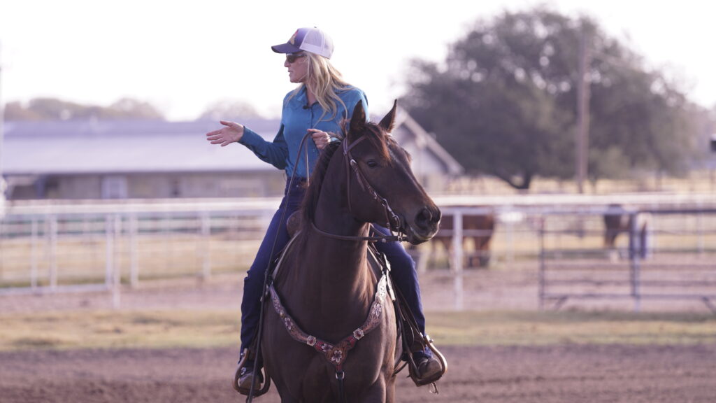 Blonde rider in a ball cap and teal shirt gestures with one hand while sitting astride a dark horse in a dirt arena