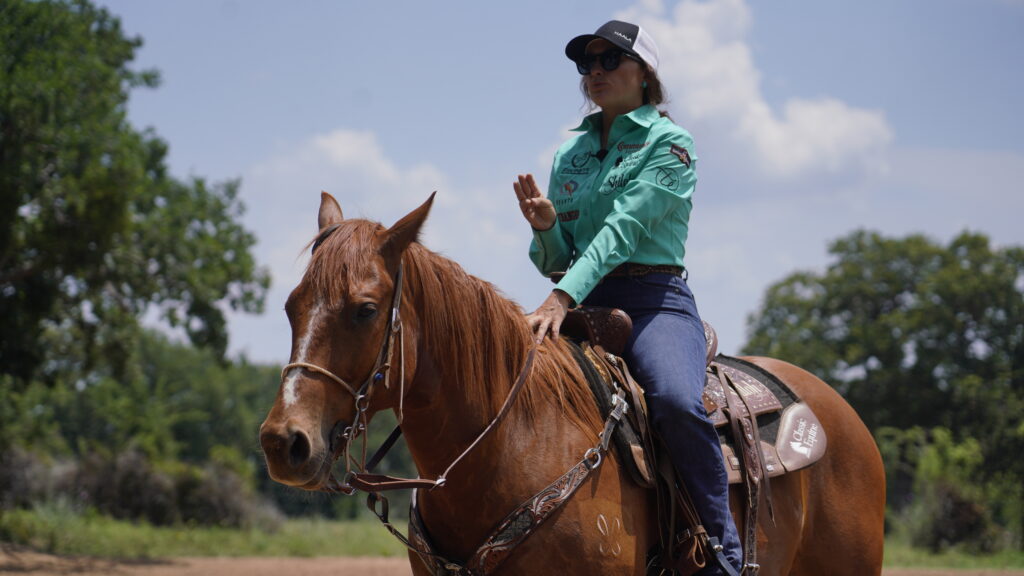 Cowgirl in turquoise shirt riding brown horse across open pasture