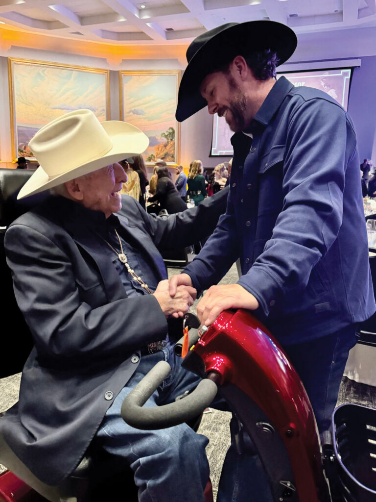 Two men in cowboy hats shake hands over a red saddle at an indoor event with spectators in the background