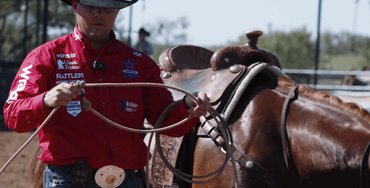 Cowboy in a red sponsor-covered shirt builds a loop with his rope while standing beside a saddled horse