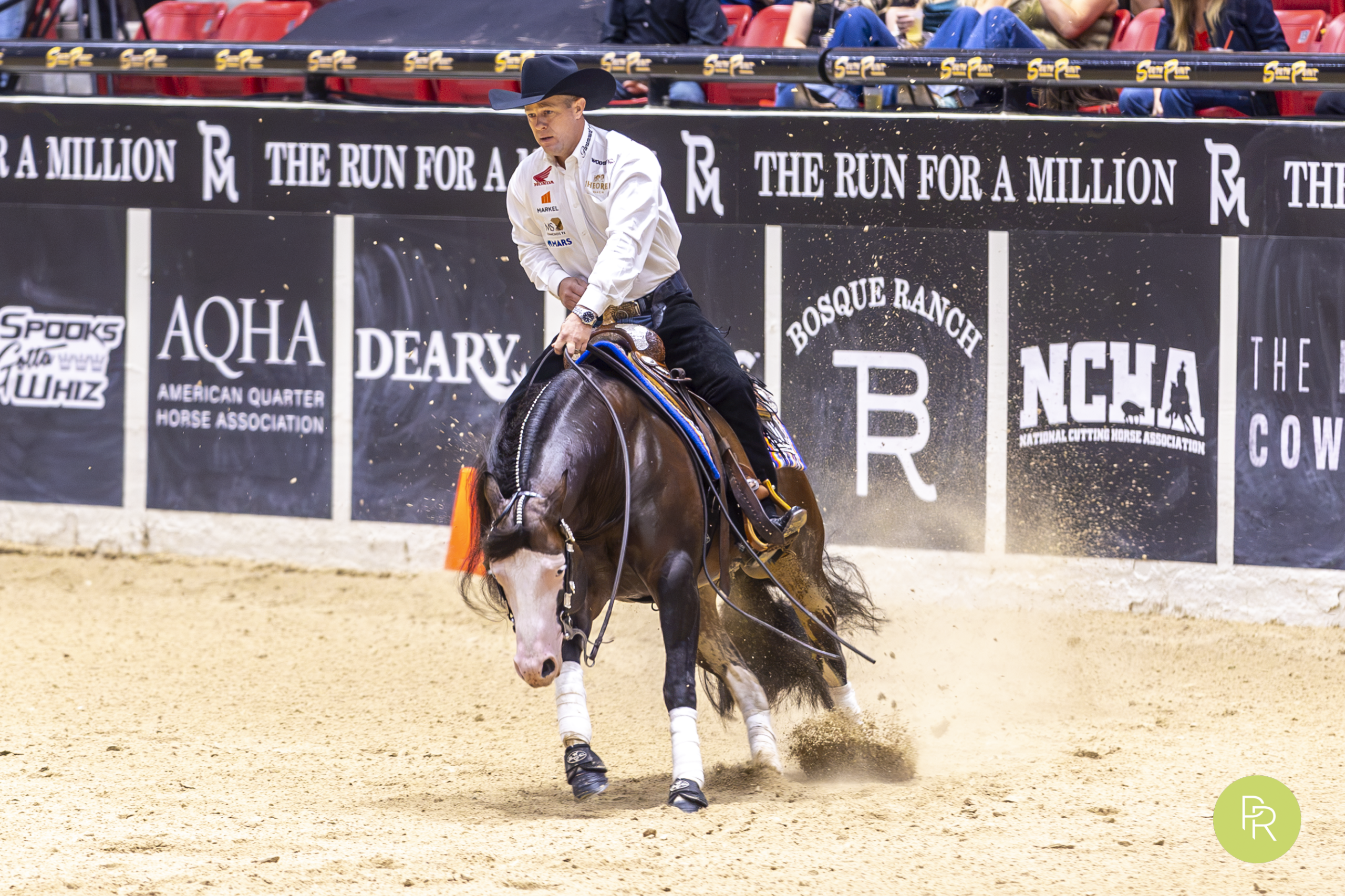 Barrel racer on a dark horse making a sharp turn around a barrel during competition at an indoor rodeo arena