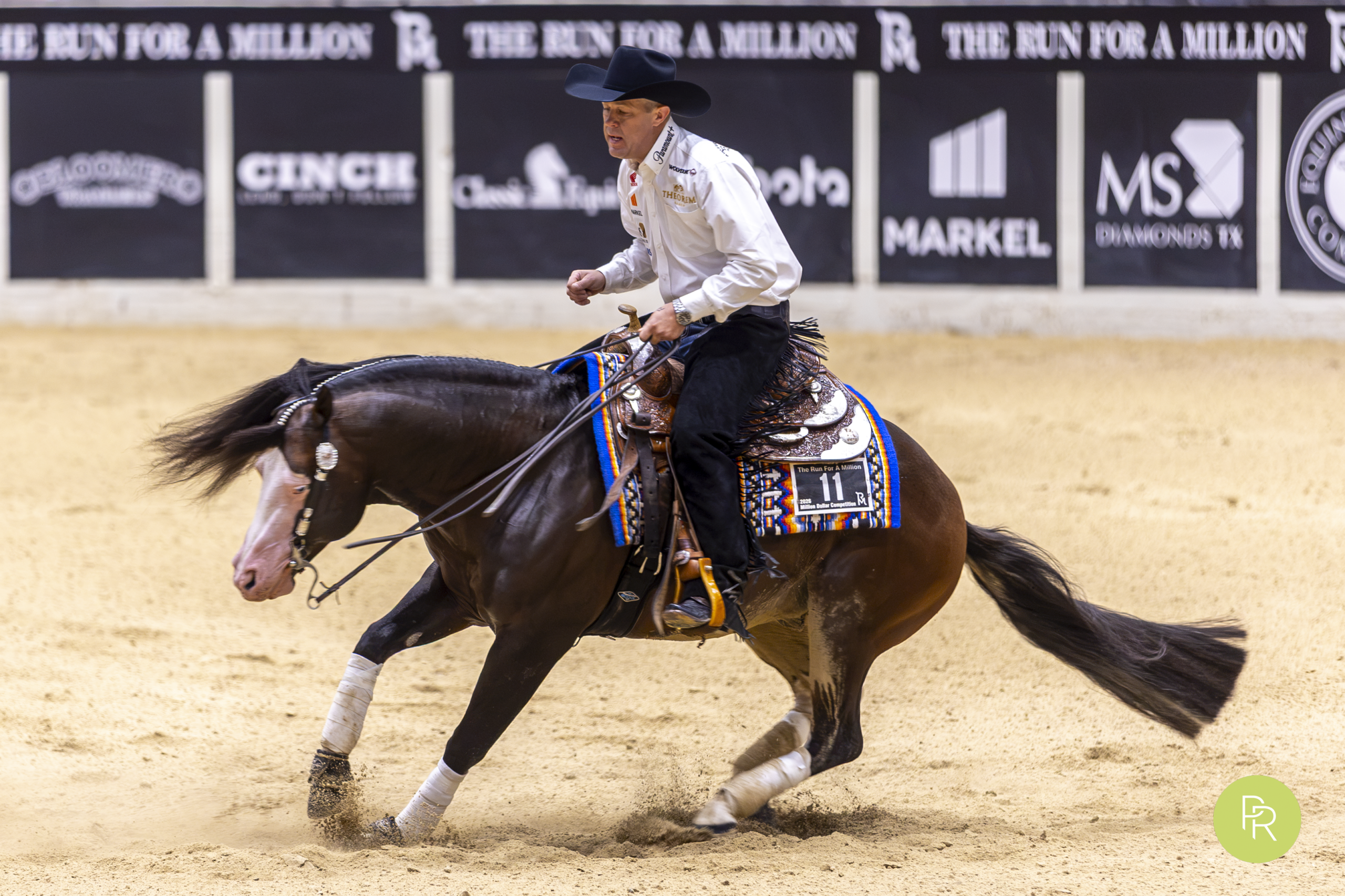 Barrel racer on dark horse turning a barrel during competition at an indoor arena with sponsor signage visible