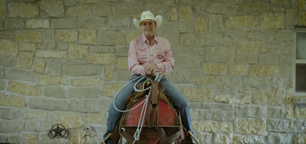 Smiling cowgirl in pink sponsor shirt and white cowboy hat sits mounted with rope in hand before a stone wall