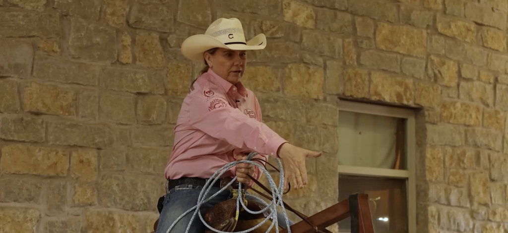 Roper in a pink shirt and straw cowboy hat holds a coiled rope while pointing and explaining beside a stone wall
