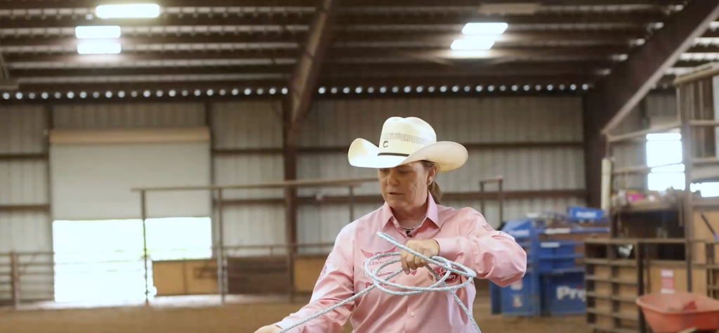 Woman in a white cowboy hat and pink western shirt coiling her rope inside an indoor arena