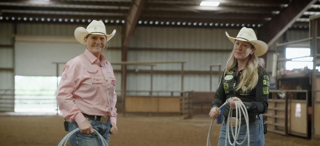 Two women in cowboy hats hold coiled ropes in a covered arena, one in a pink western shirt and one in black