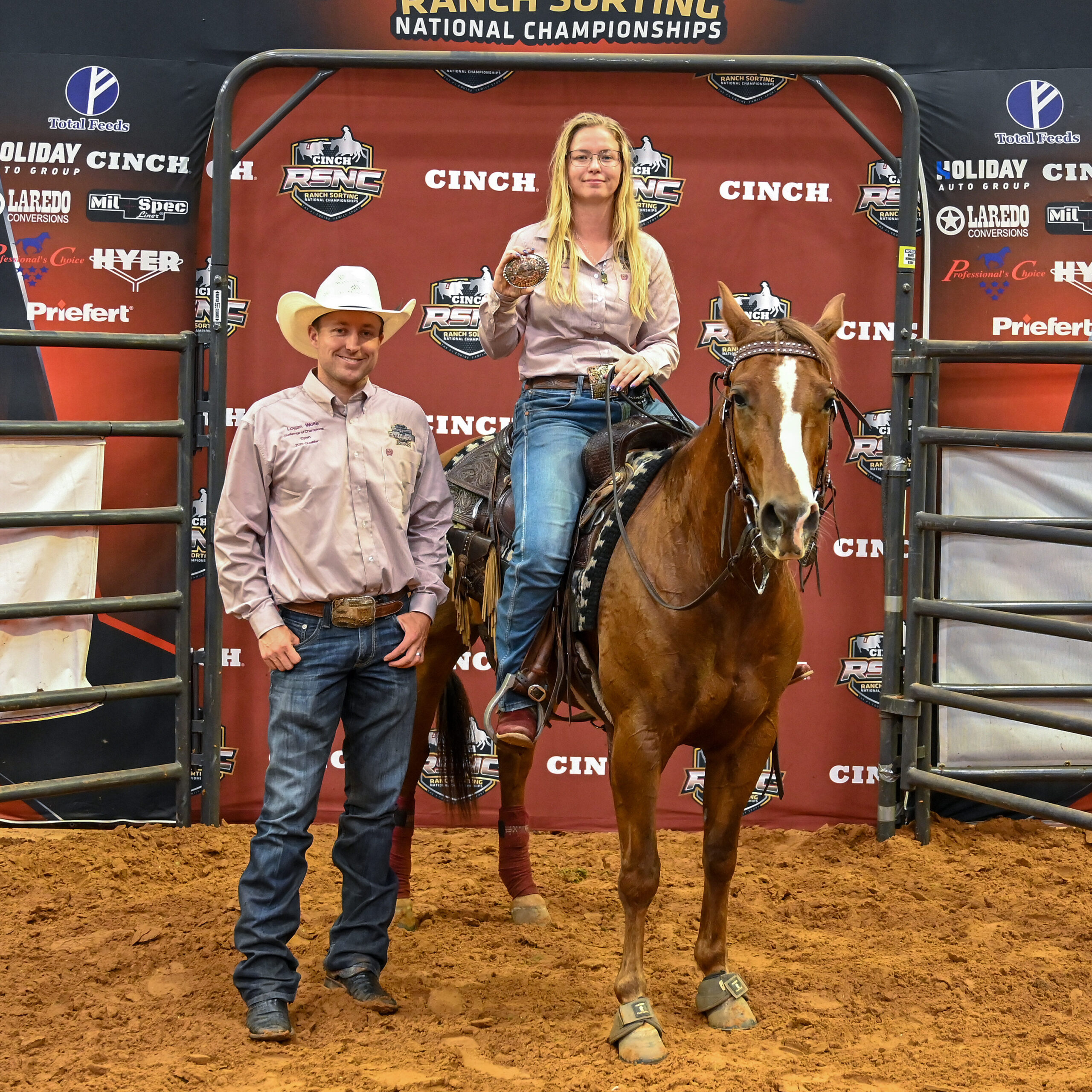 Blonde rider aboard a sorrel horse holds up a championship buckle beside a man in a white cowboy hat at the Cinch RSNC backdrop