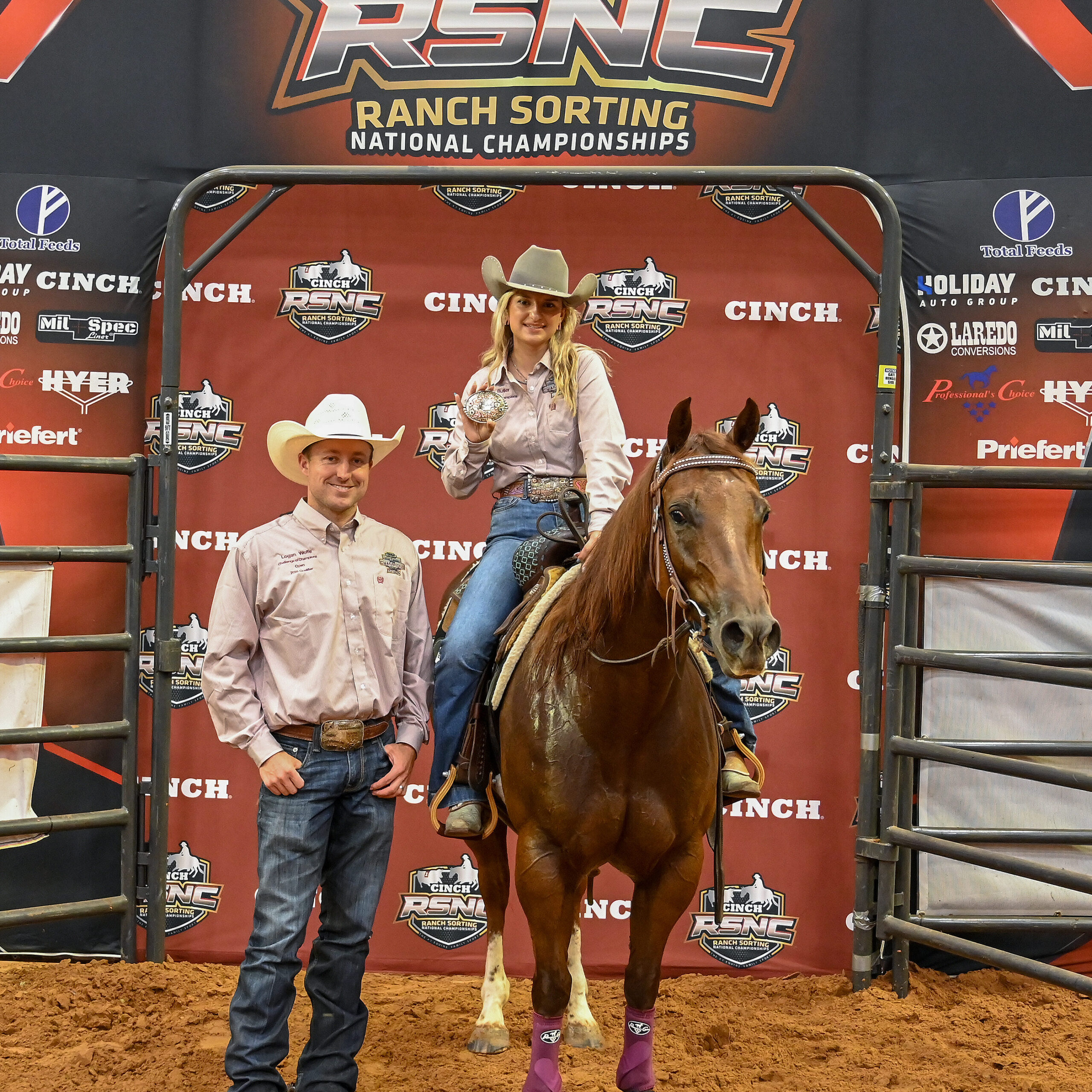 Mounted rider holds up a championship buckle beside a man in a white cowboy hat in front of a Cinch RSNC Ranch Sorting National Championships backdrop