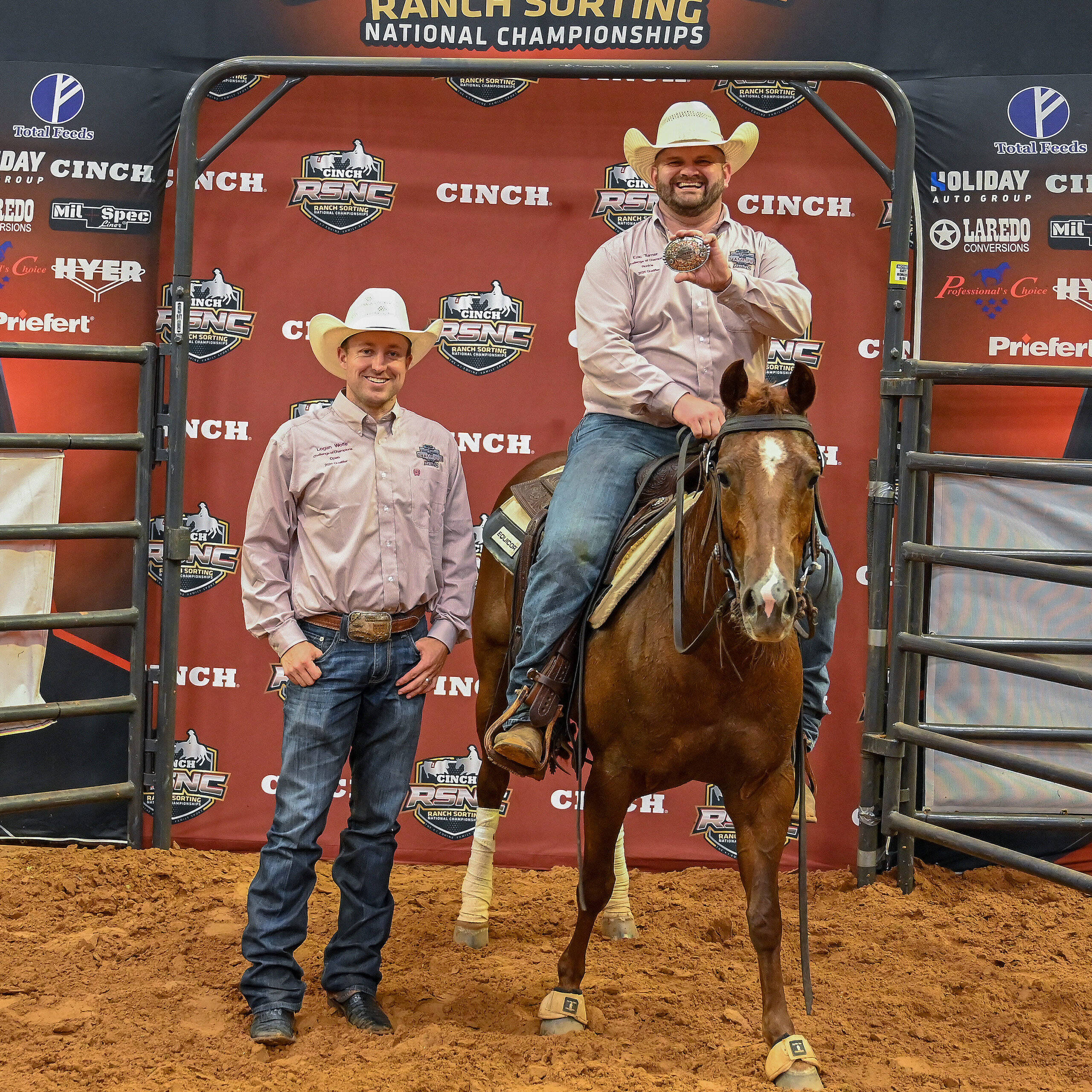 Two cowboys in matching shirts and straw hats pose for a champion photo, one mounted on a sorrel horse holding up a trophy buckle, in front of a Cinch RSNC backdrop