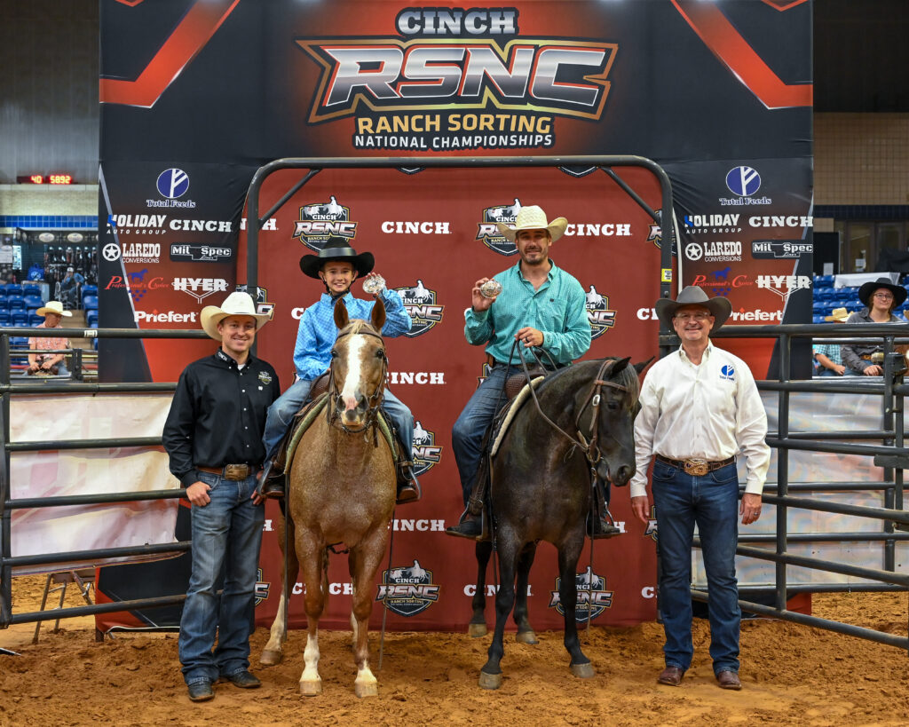 Two mounted riders hold up championship buckles beside two men on foot at the Cinch RSNC ranch sorting backdrop