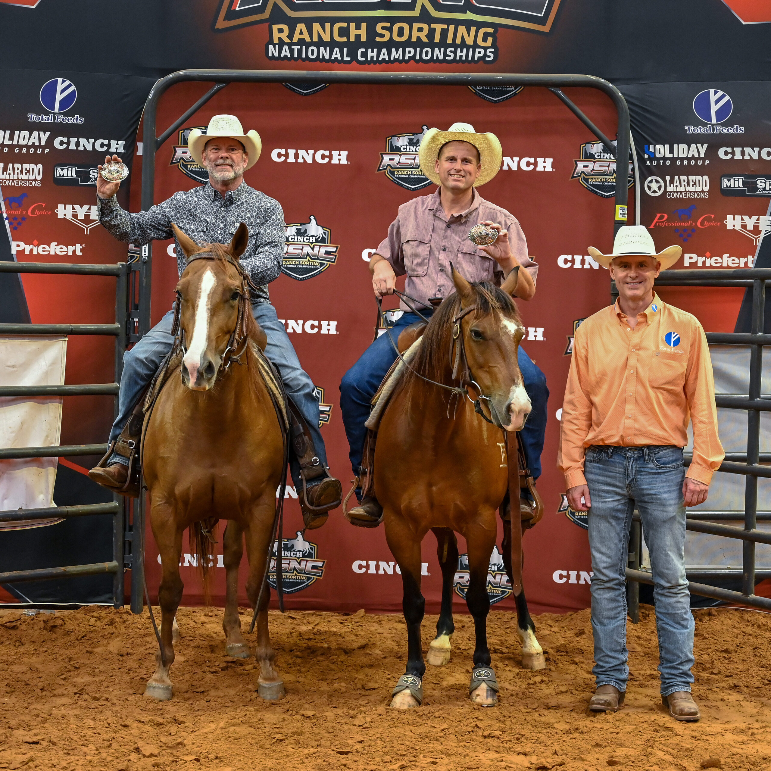 Two mounted riders hold up championship buckles beside a sponsor representative at the Cinch RSNC Ranch Sorting National Championships backdrop