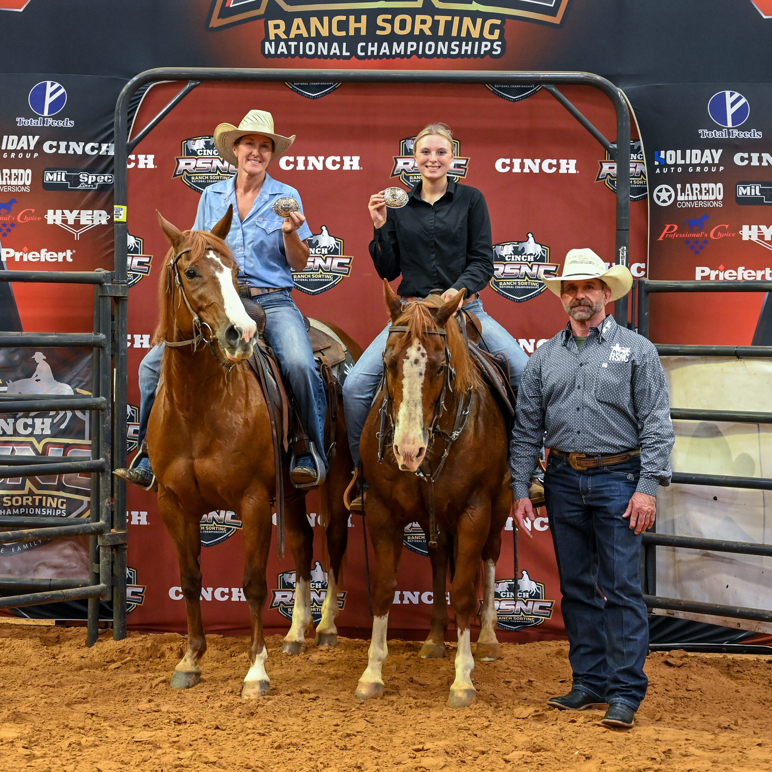Two mounted riders hold up championship buckles beside a man in a cowboy hat at the Cinch RSNC ranch sorting backdrop