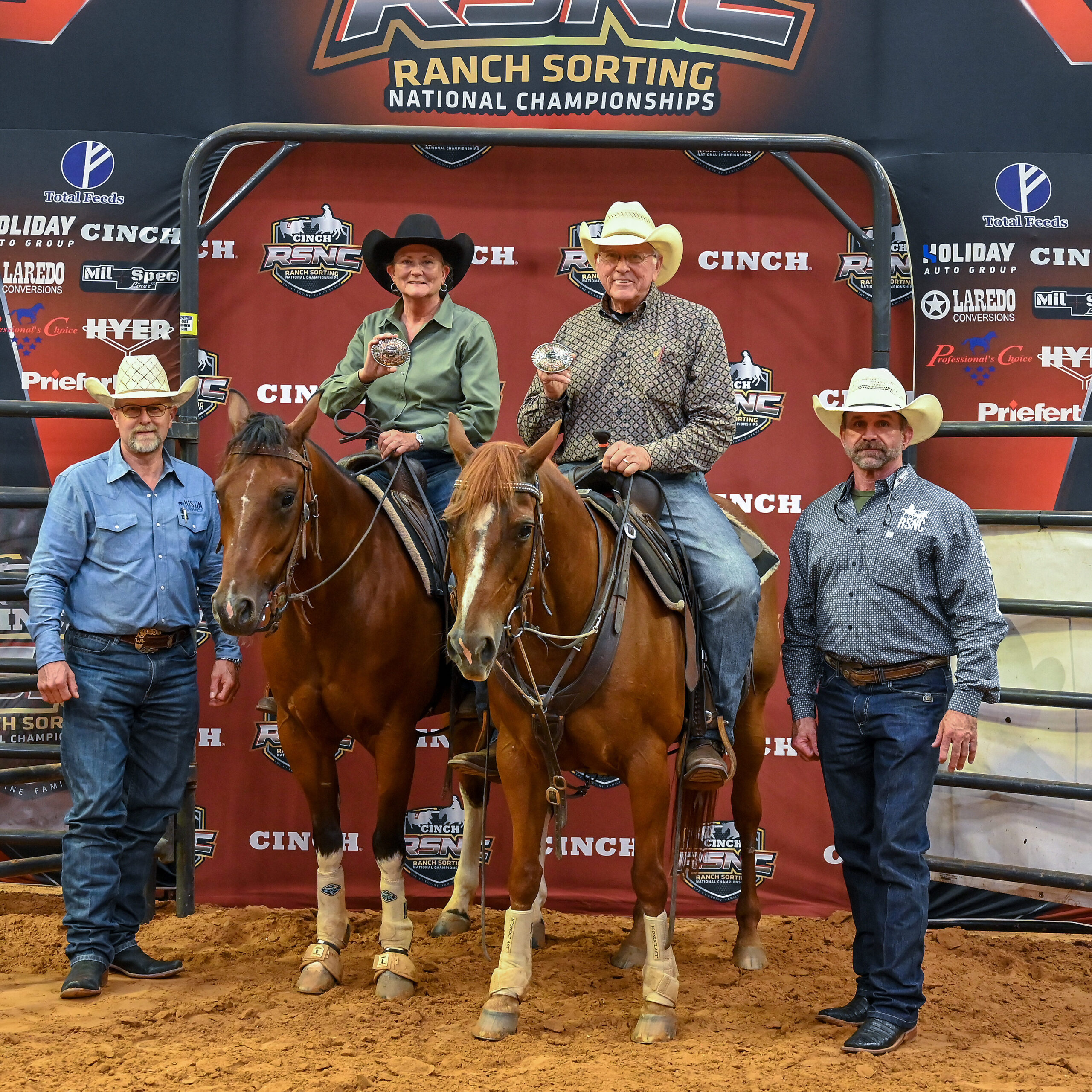 Two mounted ranch sorting partners hold up championship belt buckles while flanked by two men on foot in front of an RSNC backdrop