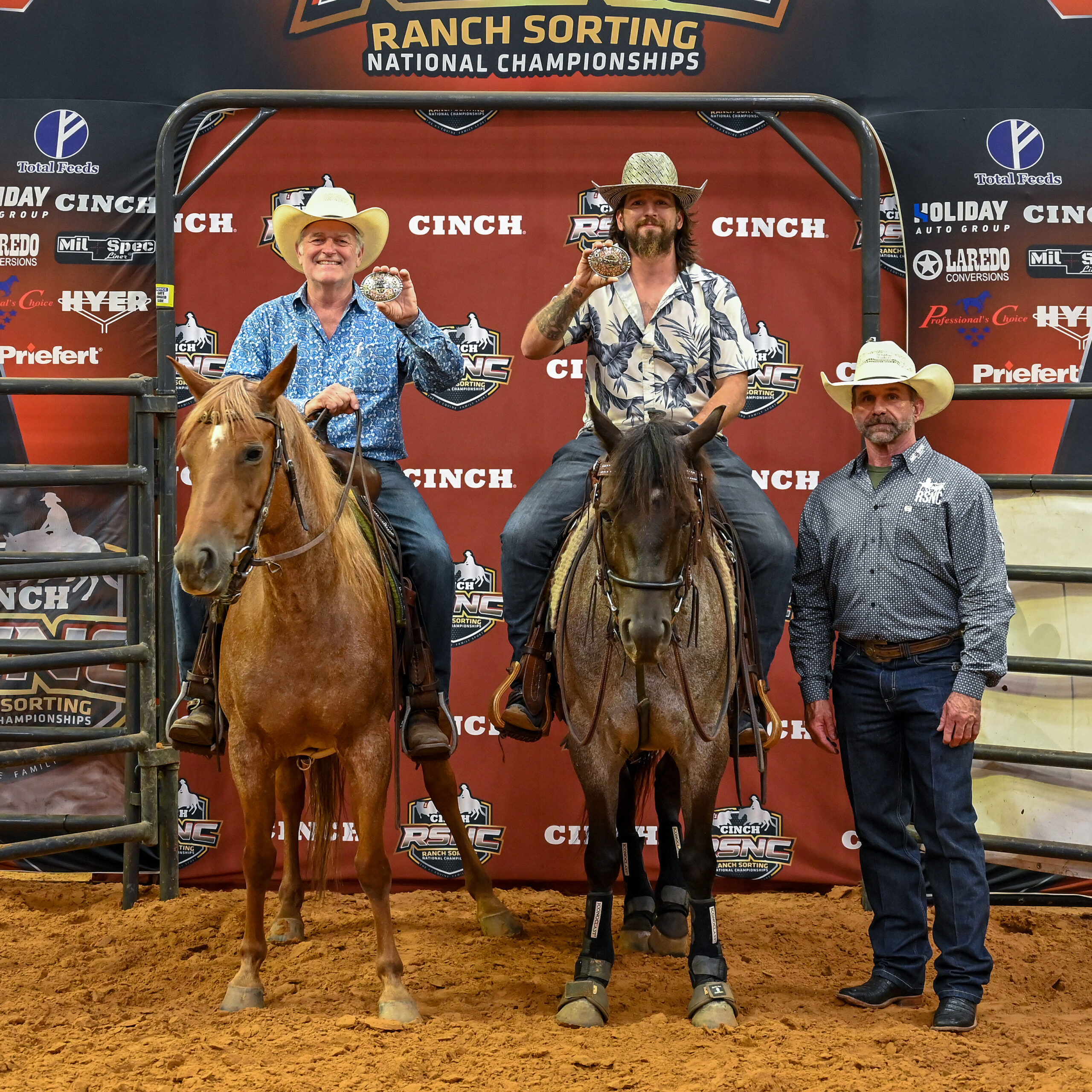 Two mounted riders hold up championship buckles beside a standing RSNC official in front of a Cinch RSNC backdrop
