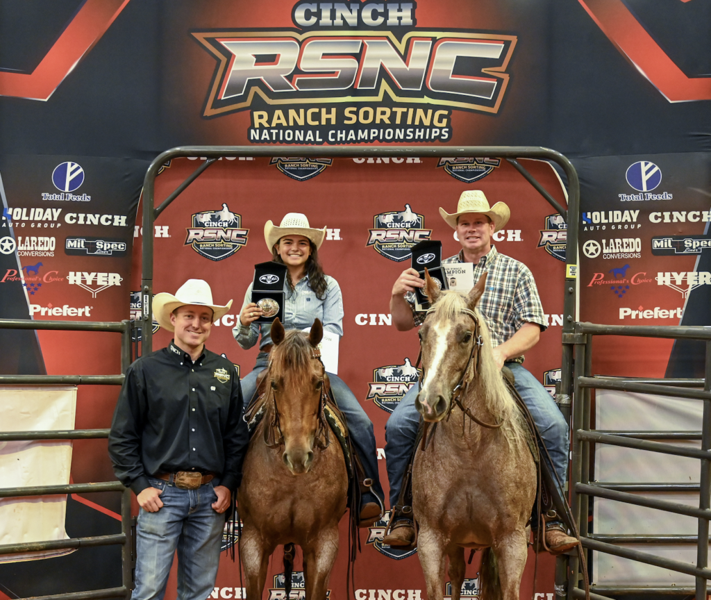 Two mounted riders in cowboy hats hold up buckle cases beside a man standing on the ground at the Cinch RSNC Ranch Sorting National Championships backdrop