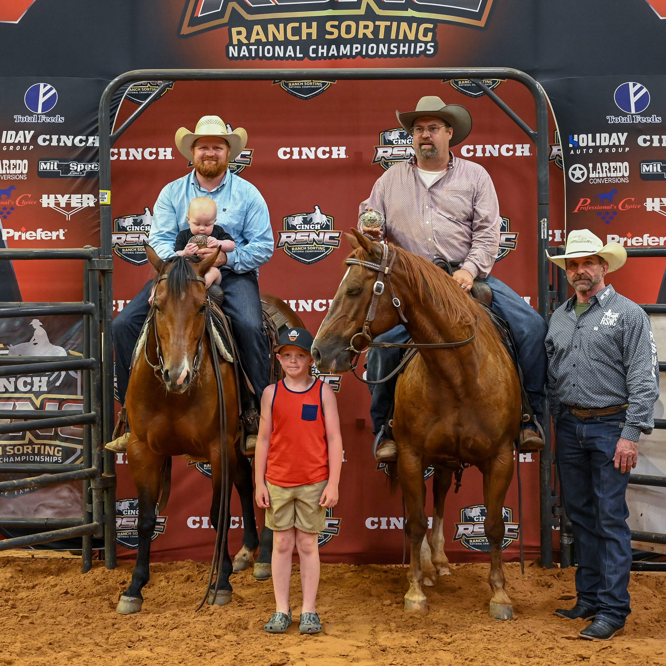 Two mounted riders hold championship buckles, one with a baby in the saddle, beside a boy and a man on foot at an RSNC ranch sorting backdrop