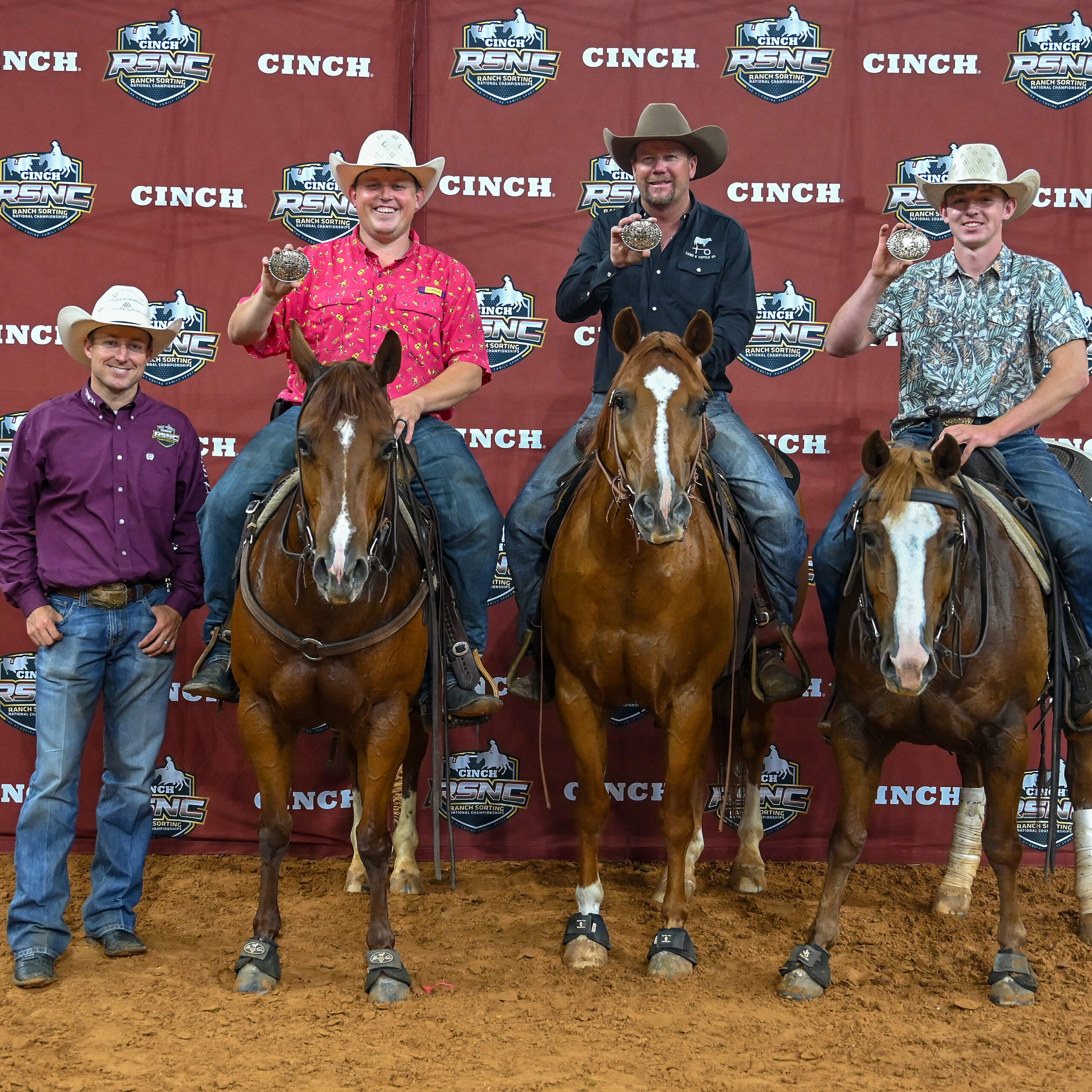 Three mounted riders hold up trophy buckles beside a man standing on the ground in front of a Cinch RSNC backdrop
