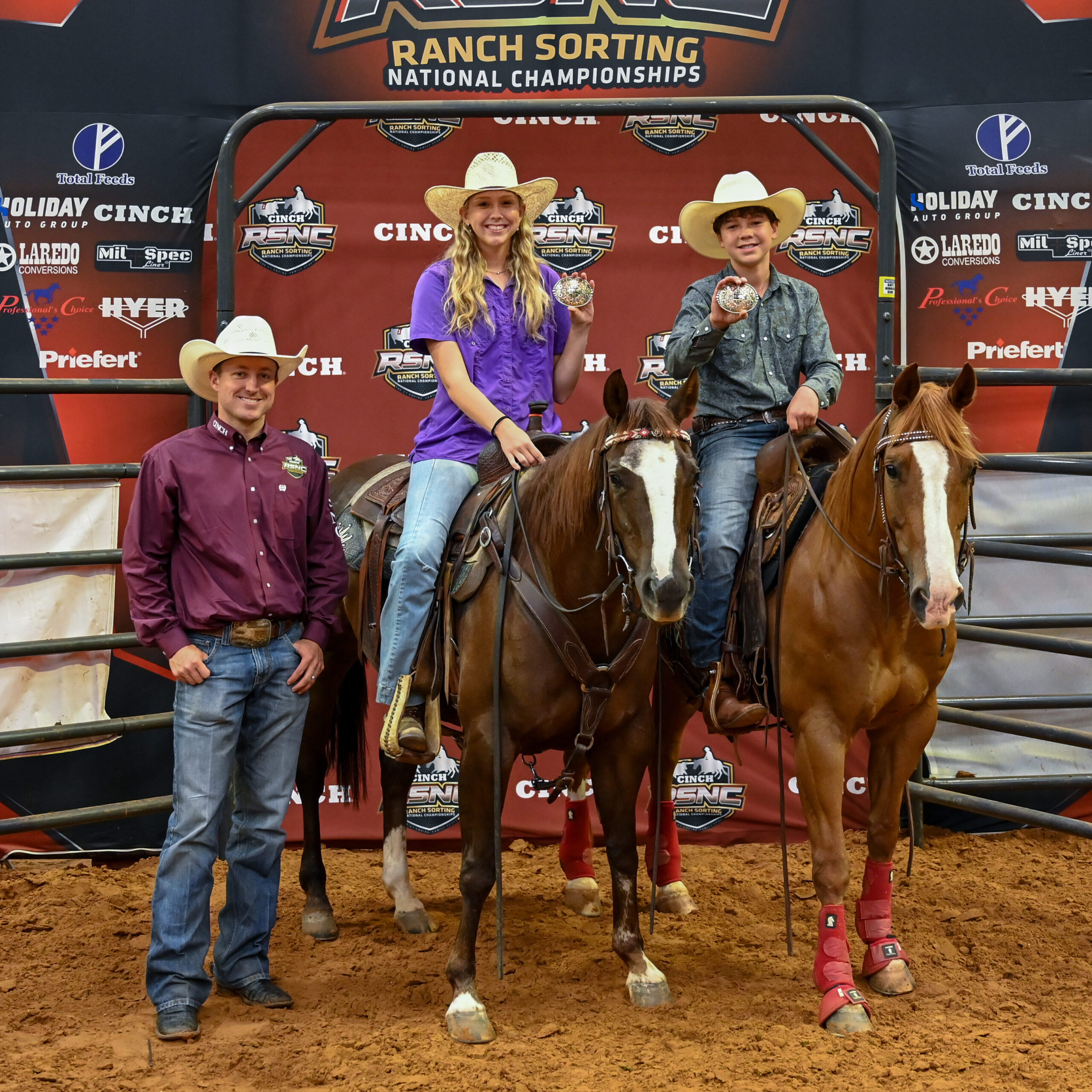 Two young riders on horseback hold up championship buckles beside a man standing on the arena dirt at the Cinch RSNC backdrop