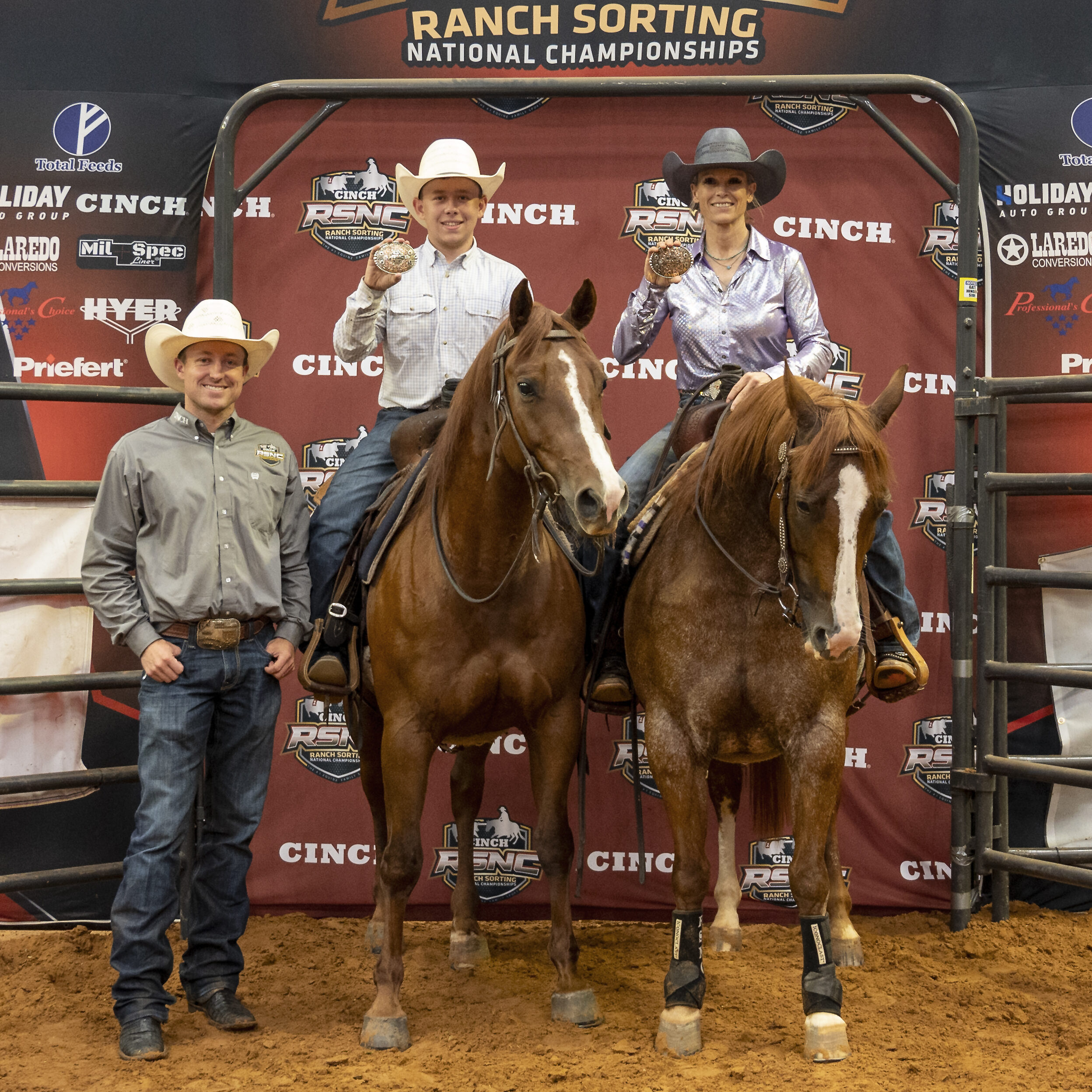 Two mounted riders hold up championship buckles beside a standing man in a cowboy hat at an RSNC ranch sorting backdrop