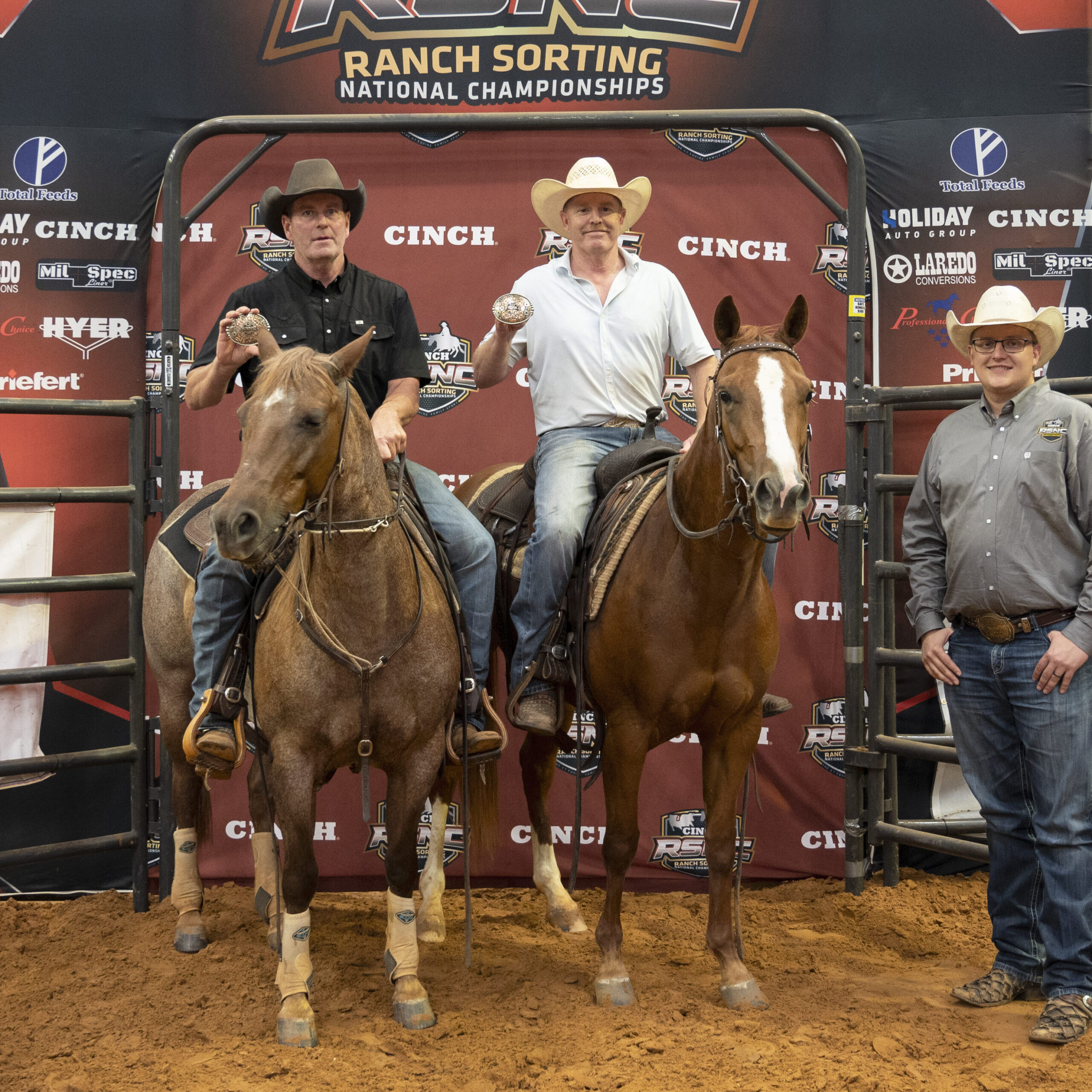 Two mounted riders hold up championship belt buckles in front of an RSNC Ranch Sorting National Championships backdrop as a man in a straw hat stands beside them