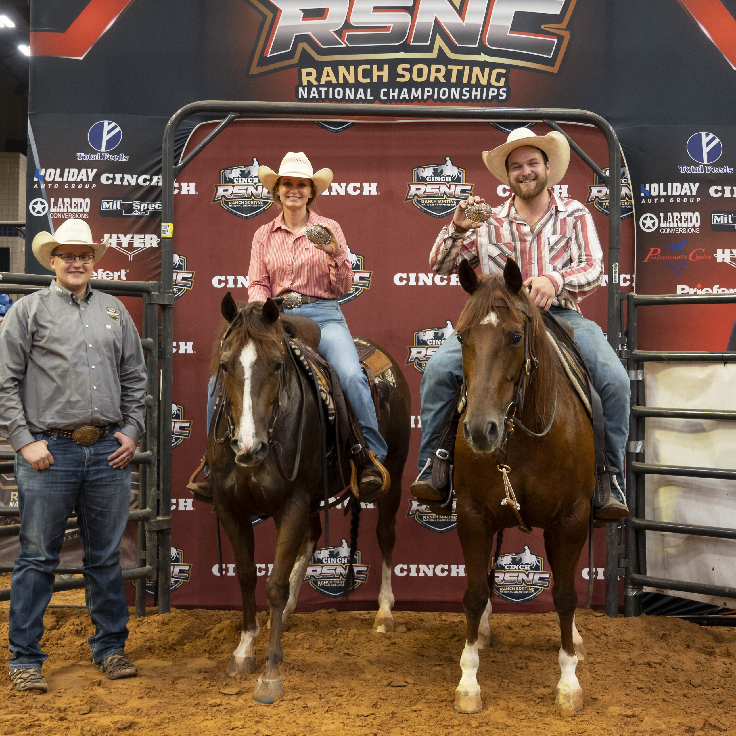 Two mounted riders hold up champion buckles beside a man on foot in front of a Cinch RSNC backdrop