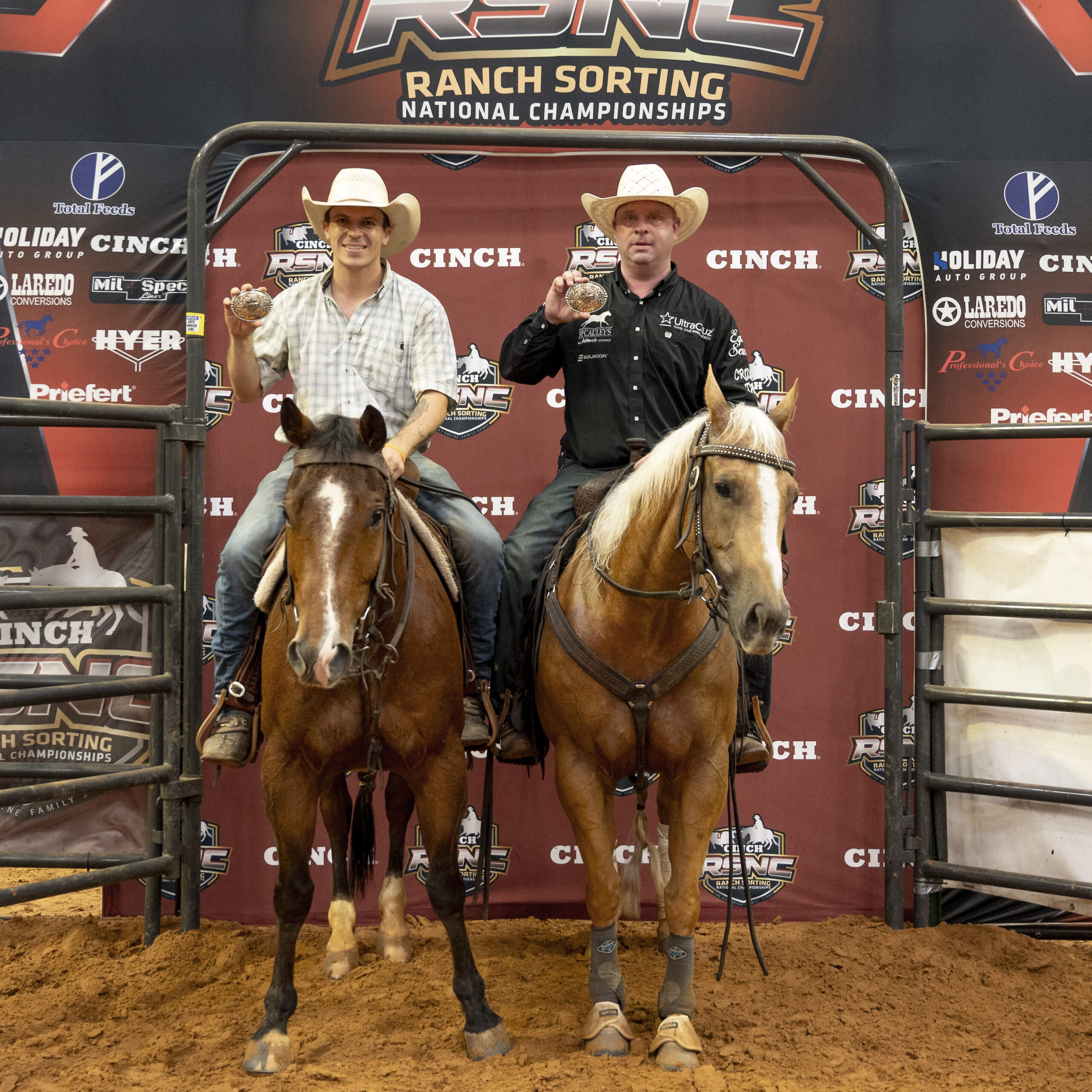 Two cowboys hold up championship belt buckles while mounted on a bay and a palomino horse in front of an RSNC Ranch Sorting National Championships backdrop