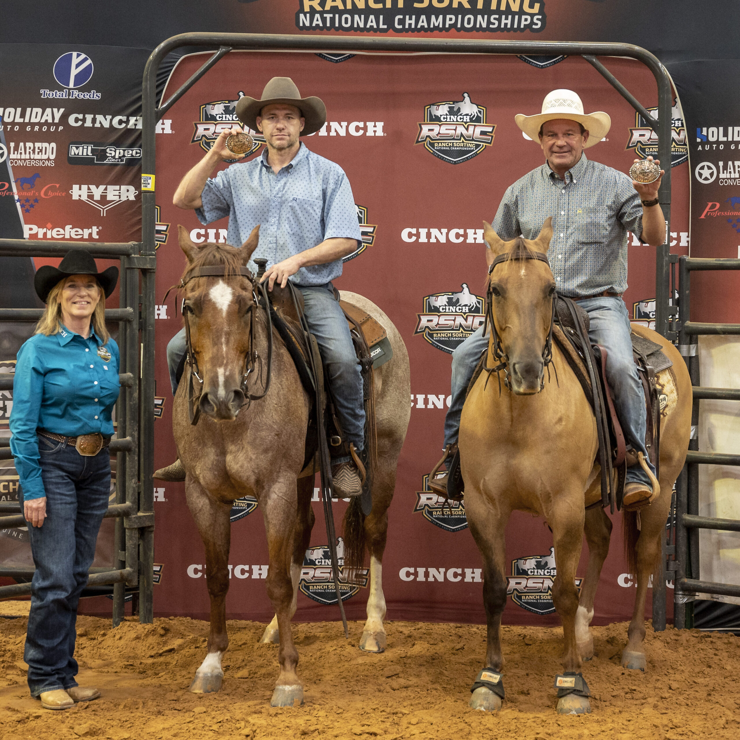 Two mounted riders hold up championship buckles beside a woman in a teal shirt in front of a Cinch RSNC backdrop