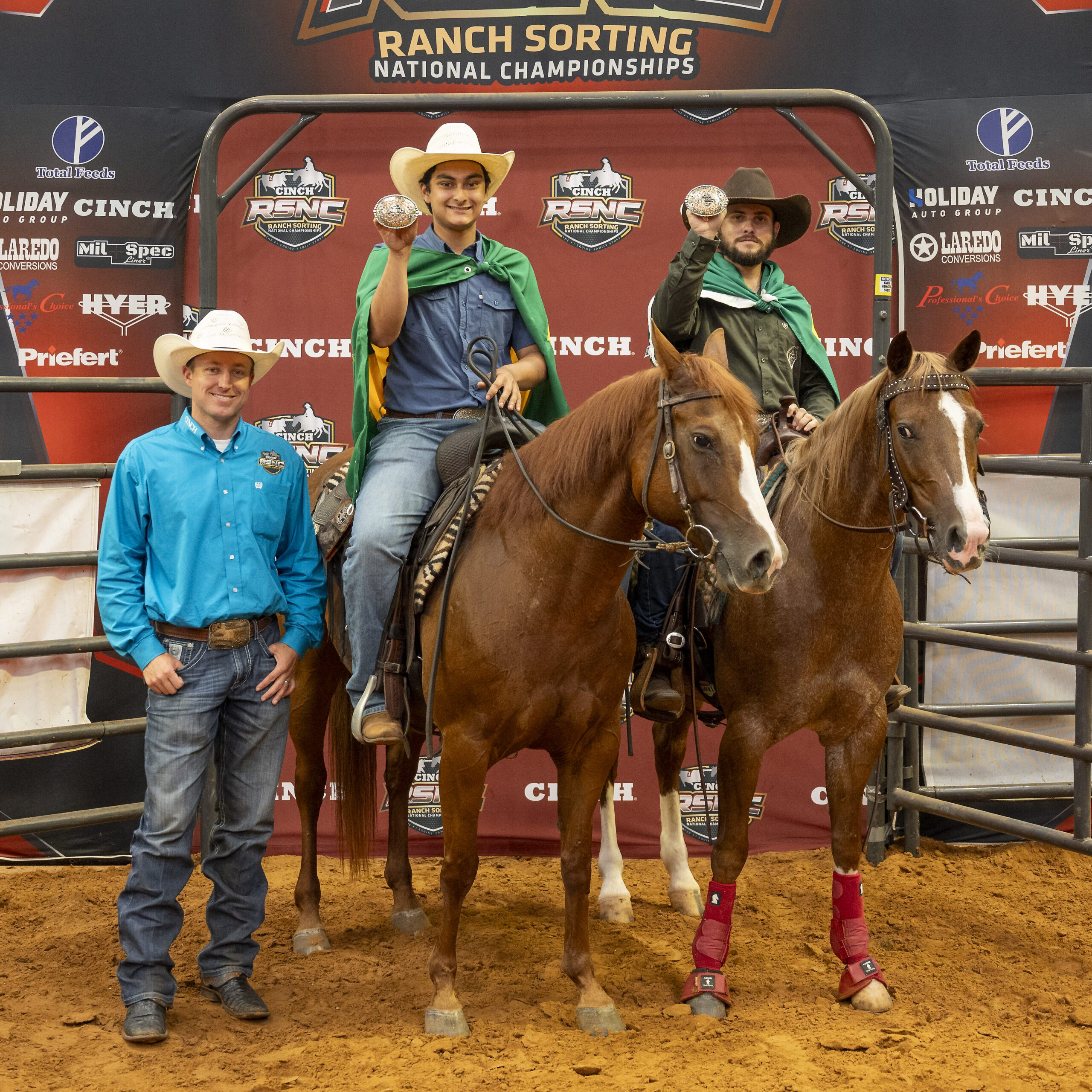 Two mounted riders wearing green capes hold up championship buckles beside a man in a turquoise shirt at the Cinch RSNC backdrop