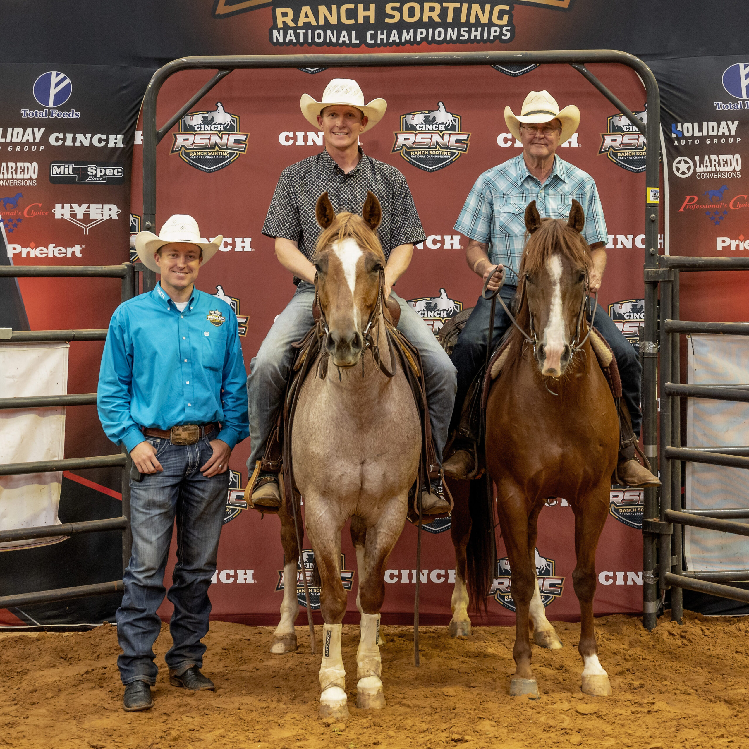 Two mounted riders in cowboy hats pose on horseback beside a standing man in a turquoise shirt at the Cinch RSNC Ranch Sorting National Championships backdrop