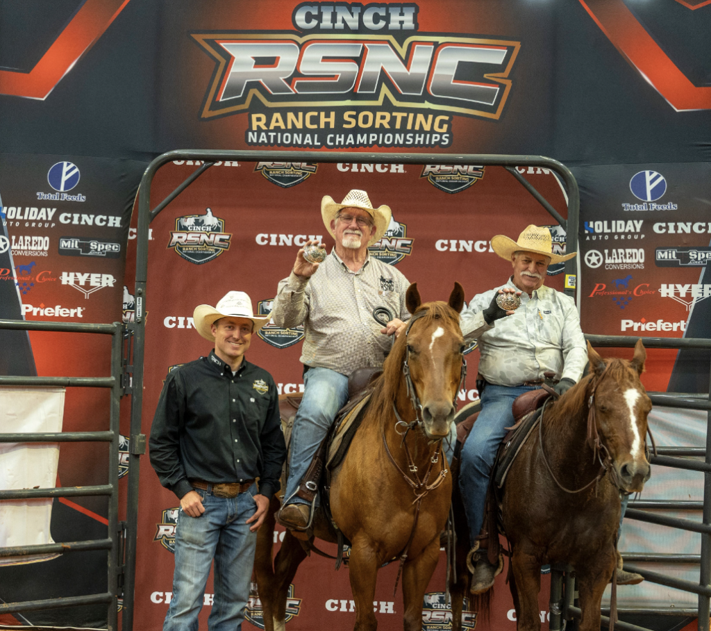 Two mounted cowboys hold up buckles beside a man standing in a black shirt at the Cinch RSNC backdrop
