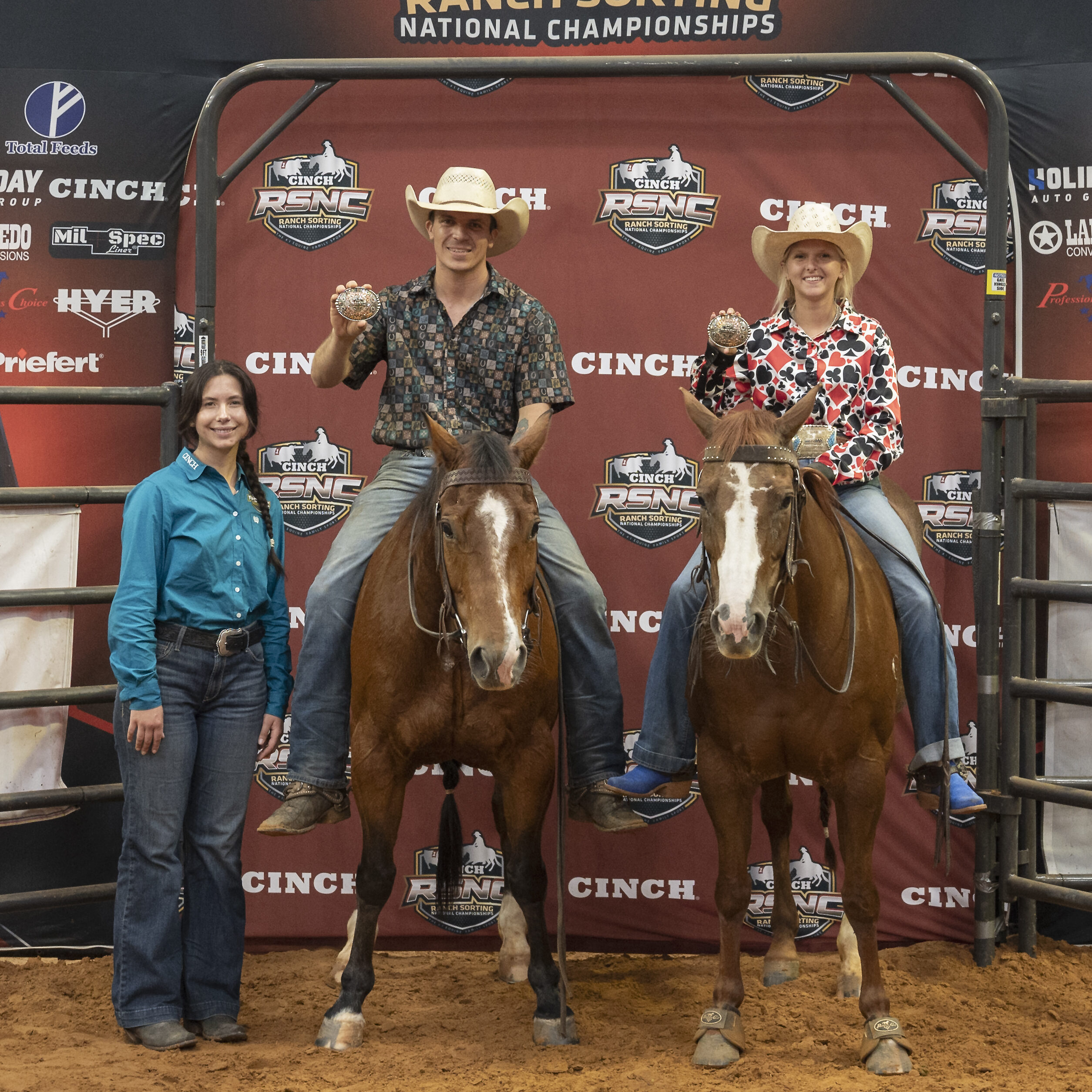 Two mounted riders hold up championship buckles beside a woman standing on the arena dirt in front of a Cinch RSNC backdrop