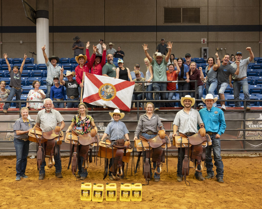 Seven winners hold trophy saddles in an arena as cheering fans wave a Florida flag in the stands behind them