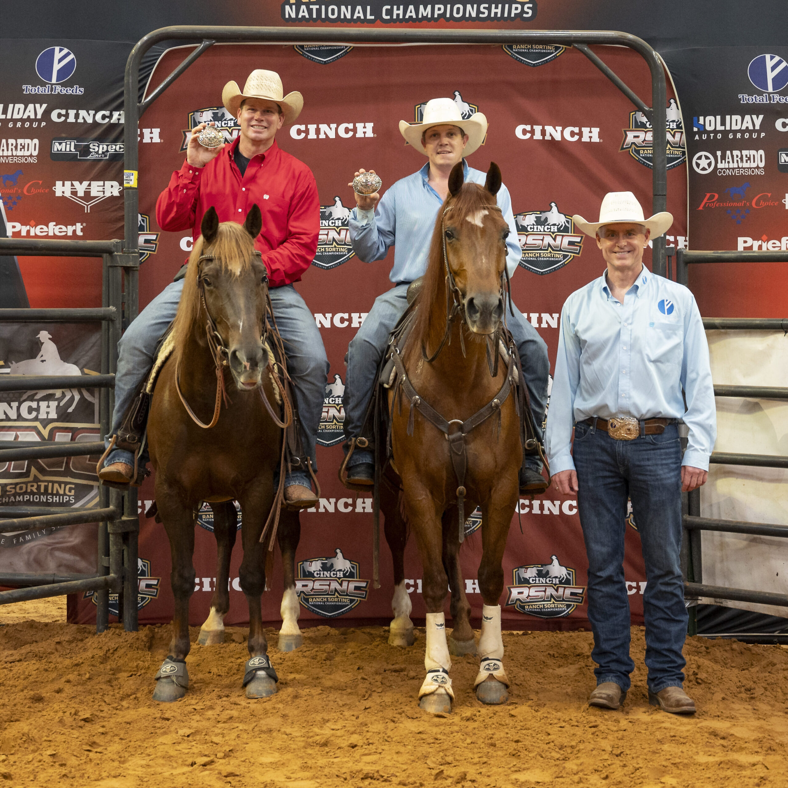 Two mounted cowboys hold up champion buckles beside a man standing in the arena at the Cinch RSNC backdrop