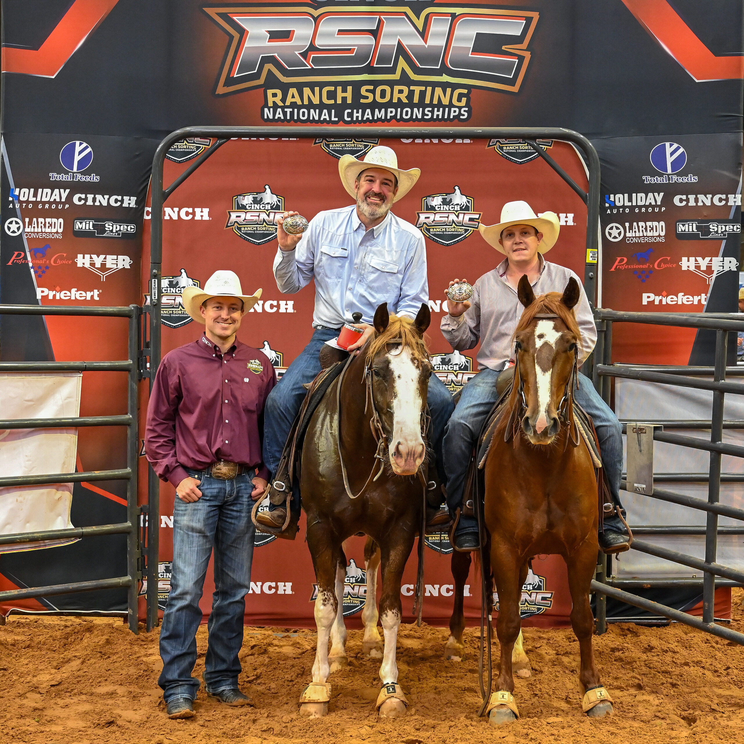 Two mounted riders hold up championship buckles beside a standing man in a maroon shirt at the Cinch RSNC backdrop