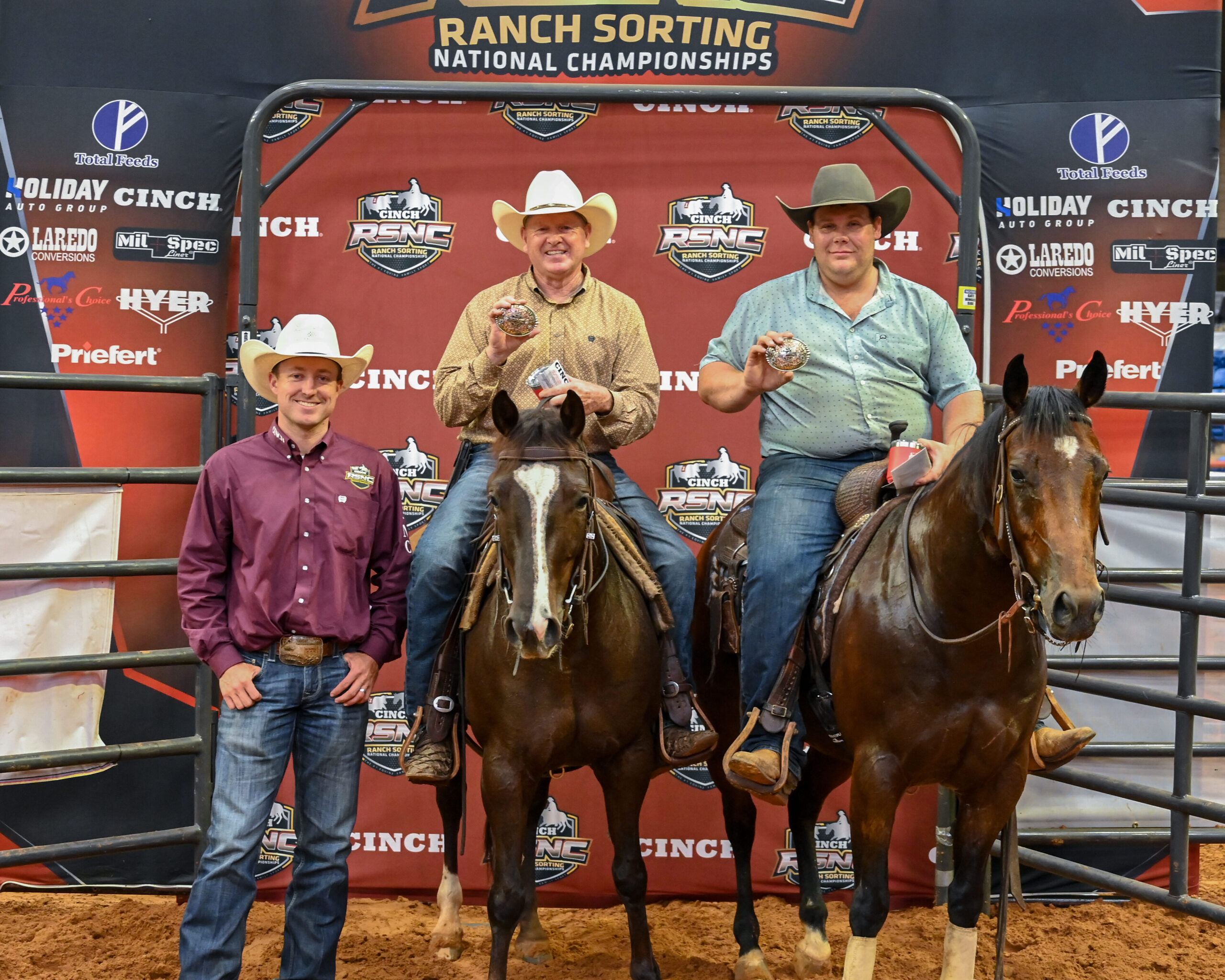 Two mounted riders hold up championship buckles beside a standing man in a cowboy hat at the Cinch RSNC Ranch Sorting National Championships backdrop